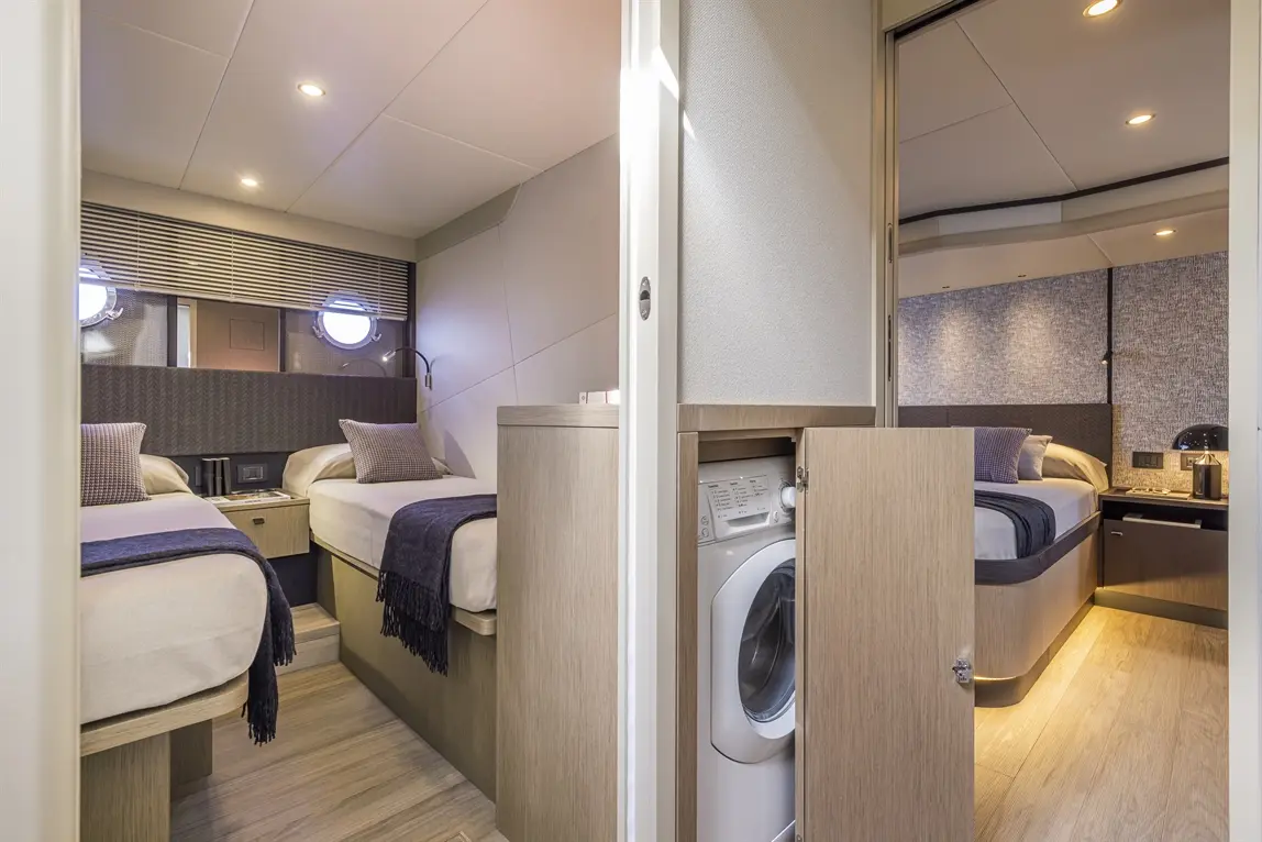 Modern boat interior featuring two single beds, a washing machine, and stylish lighting, with large porthole windows.
